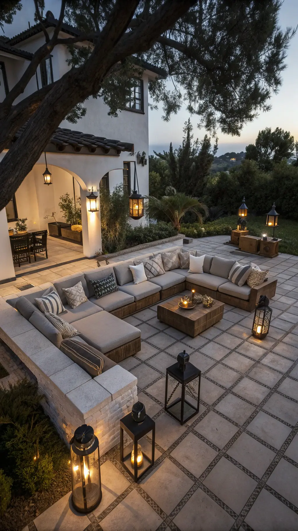 Aerial view of a moody twilight courtyard patio featuring oversized floor lanterns, a low-slung weathered grey teak sectional with ivory cushions, vintage Moroccan camel leather poufs, and black steel tables with brass vessels.