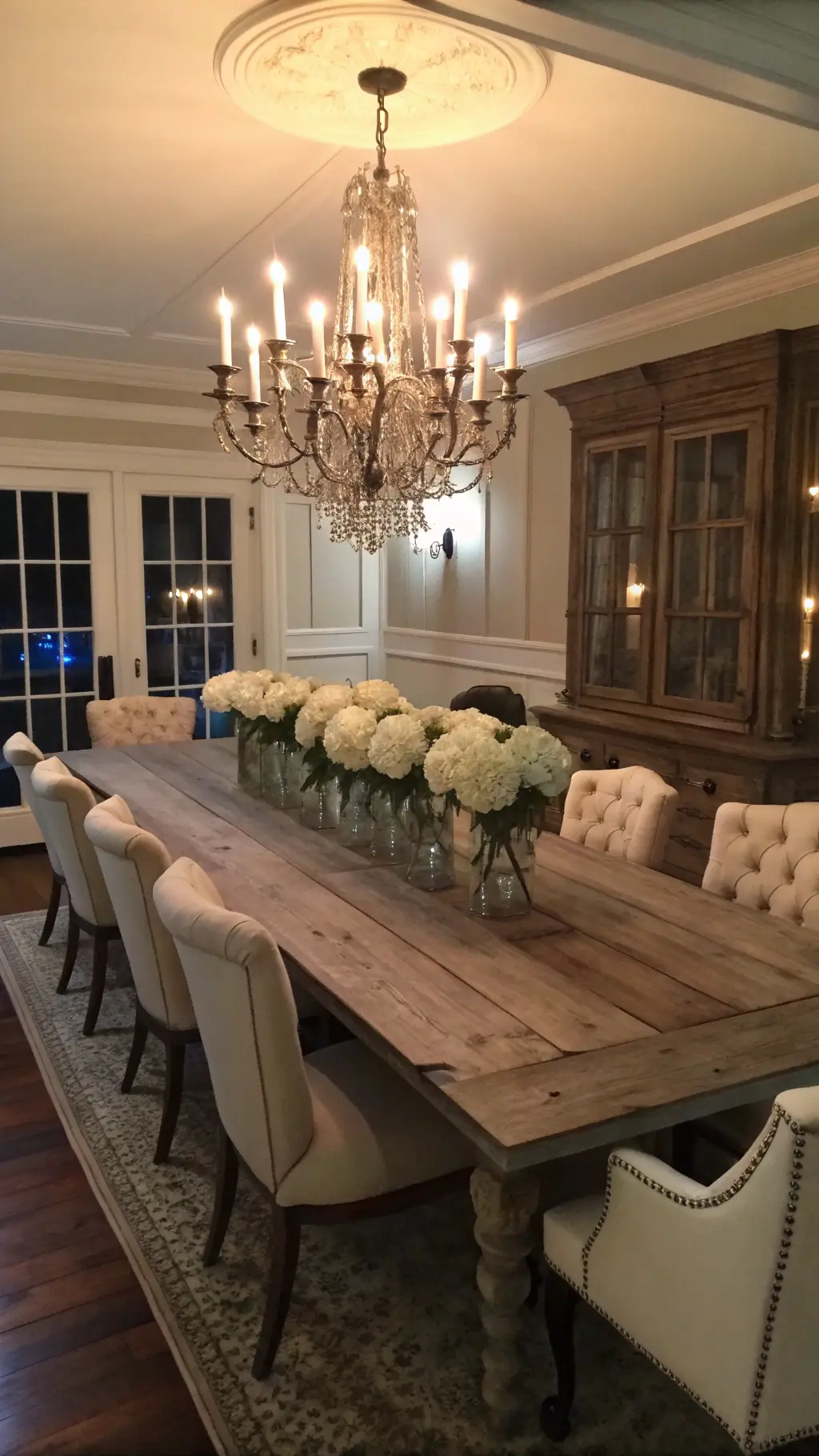 Spacious formal dining room at dusk with candlelit farmhouse table, tufted linen and painted wood chairs, crystal chandelier, blush walls with original crown molding, antiqued mirror panels and white hydrangeas in milk glass vessels.