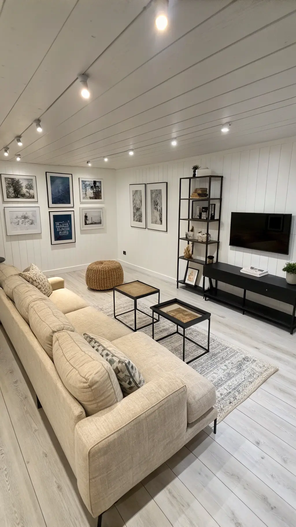 High-angle view of a minimalist Nordic den with chalk white walls, bleached pine floors, low-profile seating, black metal tables, built-in shelving, and monochrome art pieces in bright, even lighting.