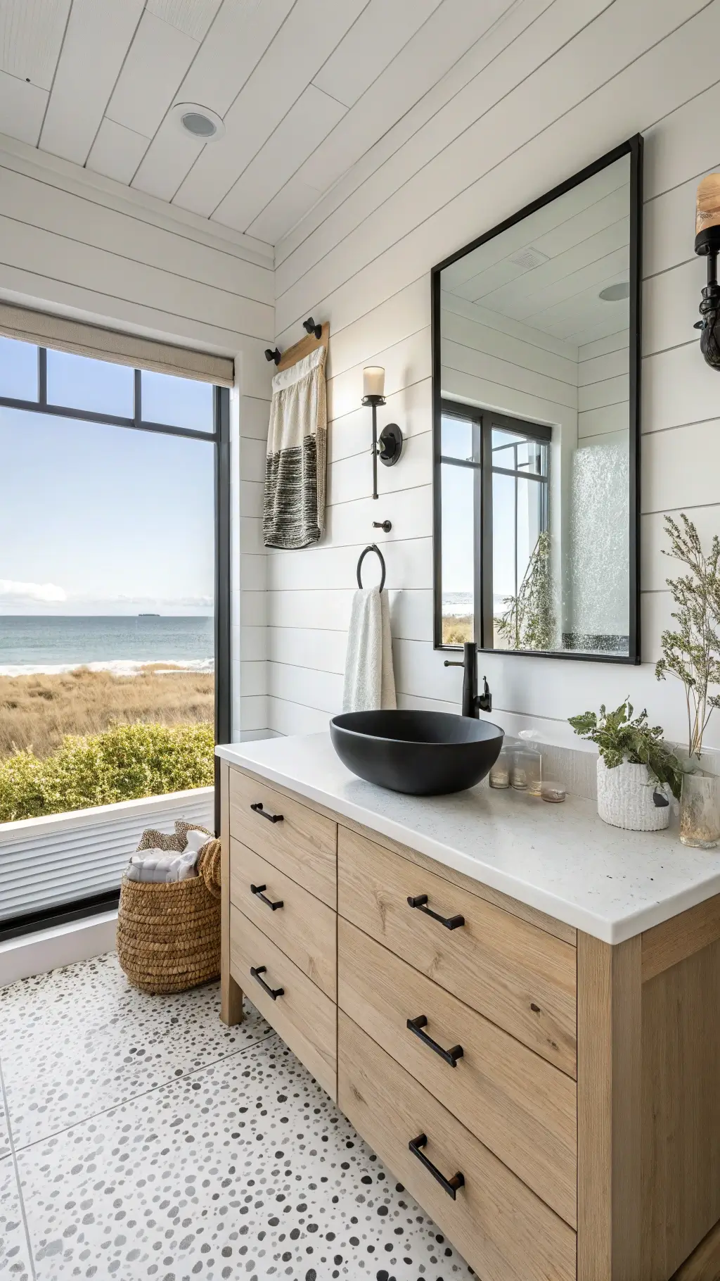 Modern coastal-inspired bathroom with white shiplap walls, black accents, floating vanity, and white terrazzo floor, decorated with coral and driftwood, overlooking the ocean